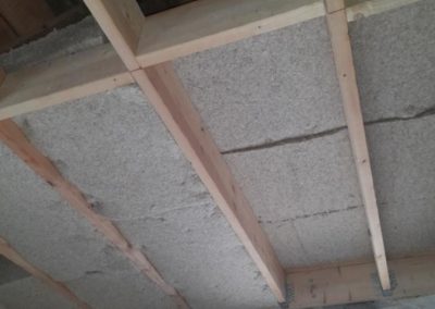 Plafond en cours d’isolation avec panneaux isolants gris insérés entre des solives en bois, au-dessus d’un mur en pierre, avec une fenêtre ouverte laissant entrer la lumière.