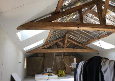 Combles en cours de rénovation avec charpente apparente en bois, isolation sous toiture avec pare-vapeur, plaques de plâtre partiellement posées, fenêtres de toit laissant entrer la lumière et matériaux de chantier au sol.